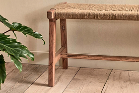 The nkuku Amhara Acacia & Jute Woven Bench sits on a light wood floor by a pale beige wall, while green houseplant leaves add natural texture and calm to the setting.