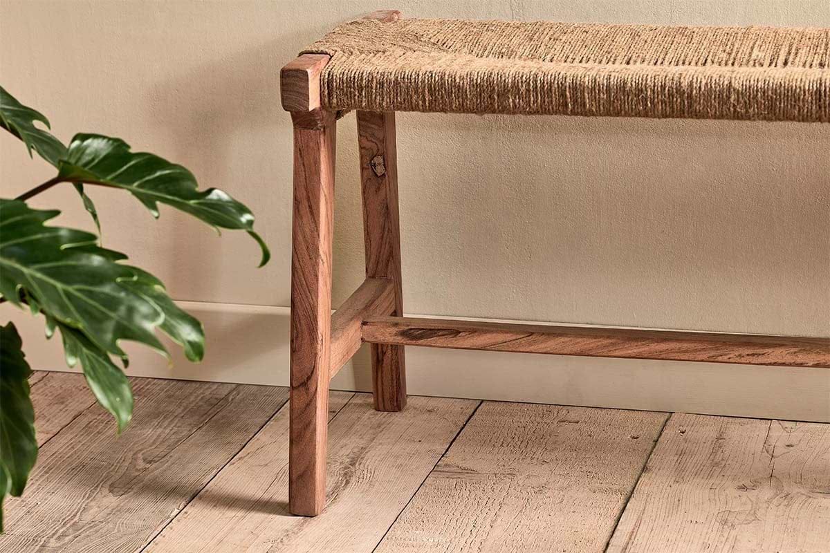 The nkuku Amhara Acacia & Jute Woven Bench sits on a light wood floor by a pale beige wall, while green houseplant leaves add natural texture and calm to the setting.