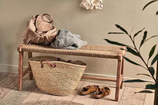 The nkuku Amhara Acacia & Jute Woven Bench holds a tan backpack and gray sweater. Below, a large straw tote and brown sandals rest on the wooden floor, with a leafy plant nearby, light walls, and a shirt hanging above.