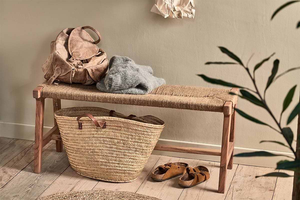The nkuku Amhara Acacia & Jute Woven Bench holds a tan backpack and gray sweater. Below, a large straw tote and brown sandals rest on the wooden floor, with a leafy plant nearby, light walls, and a shirt hanging above.