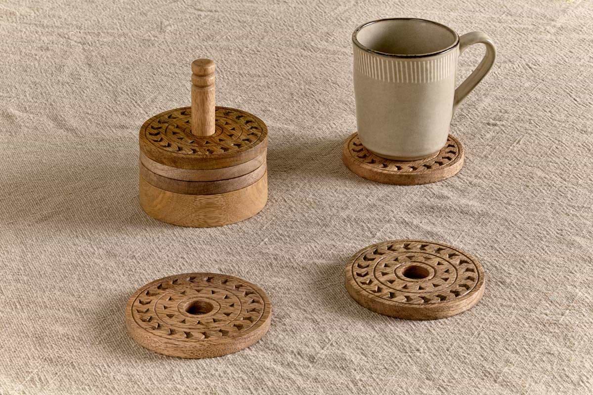 Four Anali Mango Wood Coasters by nkuku rest on a textured beige tablecloth. Three are stacked neatly in the matching holder with a convenient handle, while one supports a beige mug and two lie flat nearby. Each natural mango wood coaster features carved circular patterns and a distinctive central hole.
