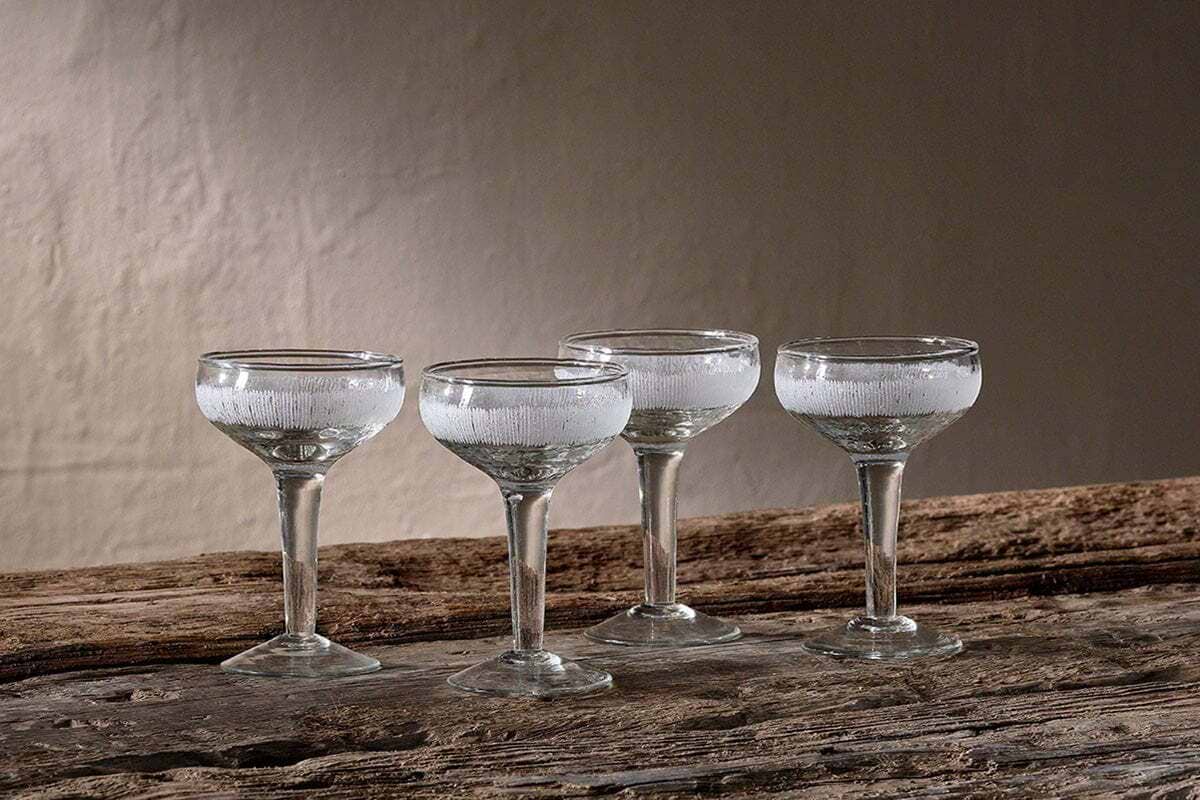 Four empty Anara Etched Champagne Glasses - Clear (Set of 4) by nkuku, with short stems, are arranged on a rustic wooden surface, their clarity and the wood’s texture highlighted by soft, neutral lighting.