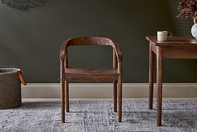 The nkuku Anbu Acacia Dining Chair in Washed Walnut, with a curved back and seat, stands on a patterned rug. To the right is an Anbu dark wood table with a mug and vase, and to the left sits a woven basket.