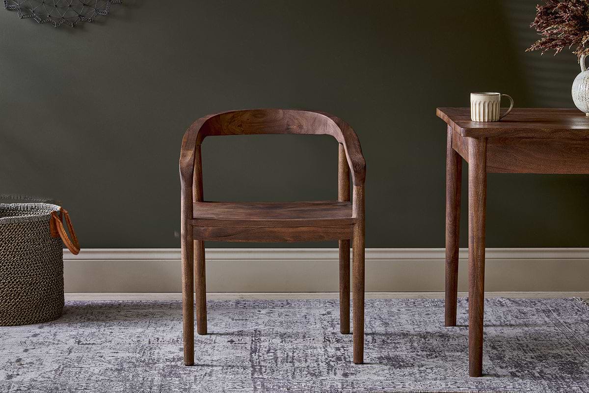 The nkuku Anbu Acacia Dining Chair in Washed Walnut, with a curved back and seat, stands on a patterned rug. To the right is an Anbu dark wood table with a mug and vase, and to the left sits a woven basket.