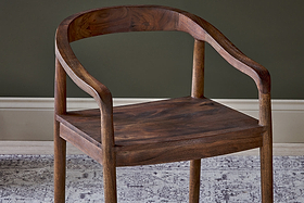 The Anbu Acacia Dining Chair by nkuku, in a washed walnut finish, features a curved backrest and armrests with smooth edges. It’s shown on a gray and white patterned rug against a dark green wall with a white baseboard.