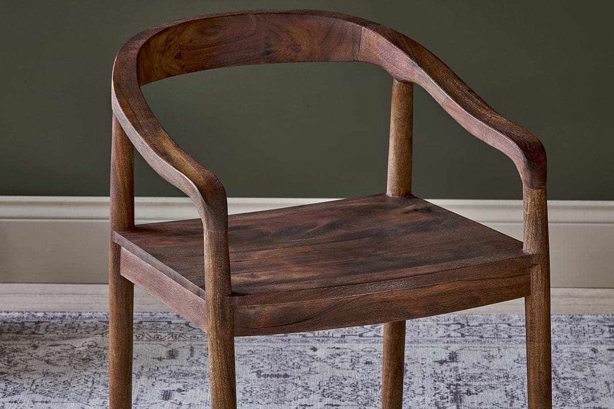 The Anbu Acacia Dining Chair by nkuku, in a washed walnut finish, features a curved backrest and armrests with smooth edges. It’s shown on a gray and white patterned rug against a dark green wall with a white baseboard.
