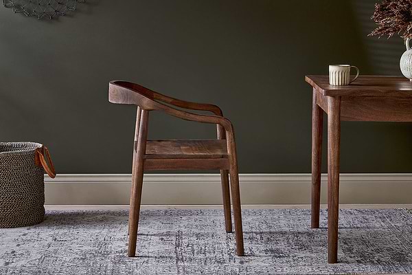 The nkuku Anbu Acacia Dining Chair in Washed Walnut sits on a patterned rug beside a dark wooden table holding a mug.