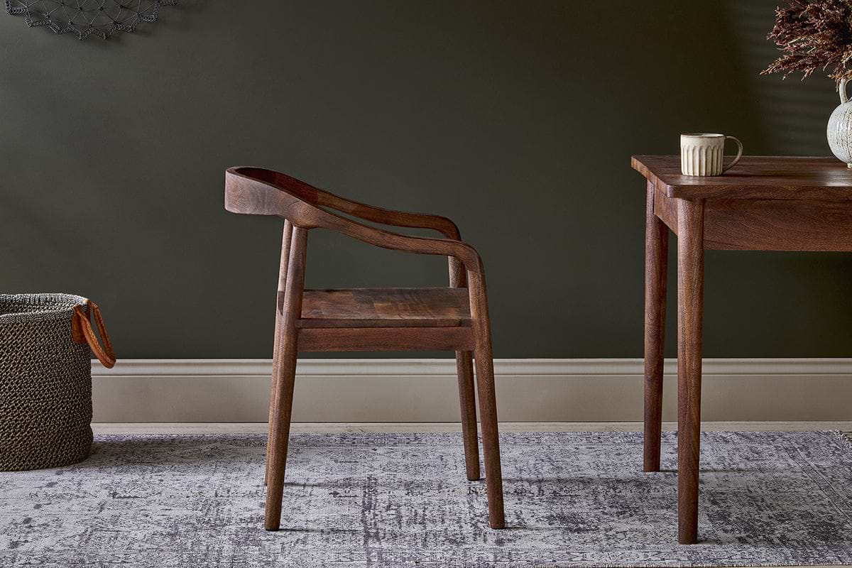 The nkuku Anbu Acacia Dining Chair in Washed Walnut sits on a patterned rug beside a dark wooden table holding a mug.