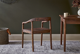 The nkuku Anbu Acacia Dining Chair in Washed Walnut sits by a matching table with a striped mug. Behind, a dark green wall, light baseboard, gray patterned rug, and a woven basket complete the scene.
