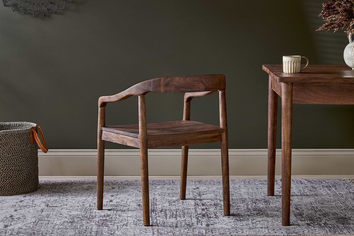 The nkuku Anbu Acacia Dining Chair in Washed Walnut sits by a matching table with a striped mug. Behind, a dark green wall, light baseboard, gray patterned rug, and a woven basket complete the scene.