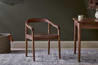The Anbu Acacia Dining Chair by nkuku, in washed walnut, features curved arms and a flat seat. It sits on a patterned rug beside a matching table with a mug and dried flowers, while a woven basket rests near the dark green wall.