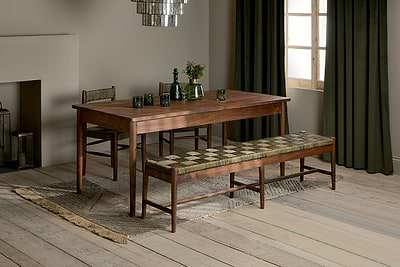 The Anbu Acacia Dining Table - Washed Walnut by nkuku sits on a rug, paired with matching chair and padded geometric bench. Green glassware, vases, emerald curtains, light wood flooring, and neutral walls complete the cozy look.
