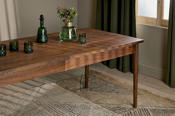 The nkuku Anbu Acacia Dining Table in Washed Walnut displays three green glass tumblers, a green bottle, and a clear vase with flowers on a beige patterned rug near green curtains and a sunlit window.