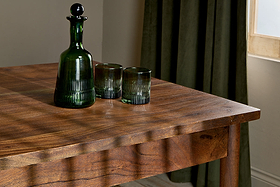 A dark green glass decanter with a round stopper and two matching tumblers sit on the nkuku Anbu Acacia Dining Table in Washed Walnut, sunlight casting soft shadows. Behind the table, a green curtain frames a sunlit window.