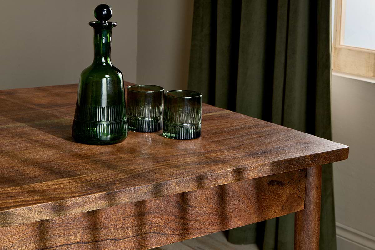 A dark green glass decanter with a round stopper and two matching tumblers sit on the nkuku Anbu Acacia Dining Table in Washed Walnut, sunlight casting soft shadows. Behind the table, a green curtain frames a sunlit window.