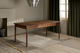 The Anbu Acacia Dining Table by nkuku, finished in washed walnut with tapered legs, stands on a textured beige rug in a sunlit room featuring wooden floors, olive green curtains, modern chandelier, and neutral minimalist walls.