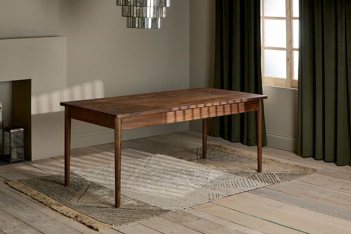 The Anbu Acacia Dining Table by nkuku, finished in washed walnut with tapered legs, stands on a textured beige rug in a sunlit room featuring wooden floors, olive green curtains, modern chandelier, and neutral minimalist walls.