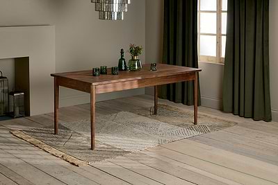 The nkuku Anbu Acacia Dining Table in Washed Walnut sits on a patterned rug, topped with three green glasses, a green bottle, and a vase of flowers. Behind it, dark green curtains frame the window.