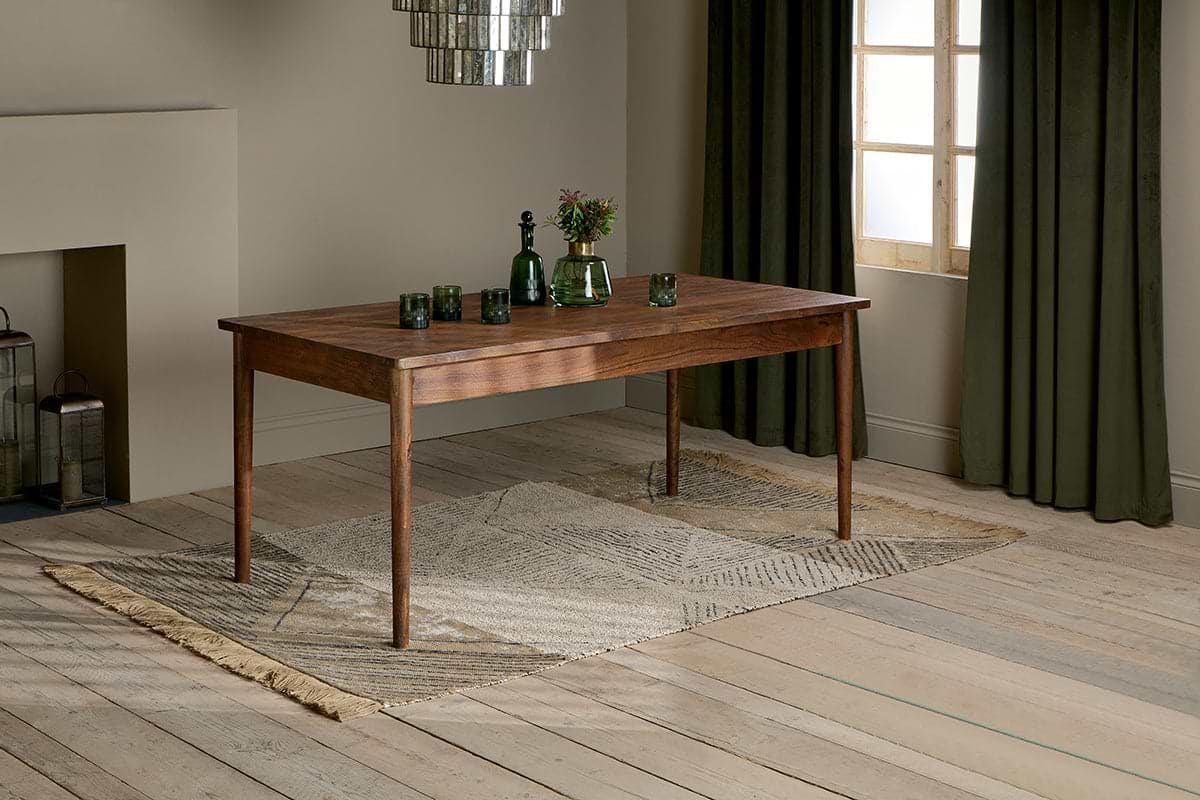 The nkuku Anbu Acacia Dining Table in Washed Walnut sits on a patterned rug, topped with three green glasses, a green bottle, and a vase of flowers. Behind it, dark green curtains frame the window.