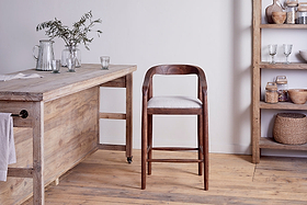 An nkuku Anbu Acacia Upholstered Counter Chair – Washed Walnut with a white cushion sits in a rustic kitchen with light wooden floors, an acacia table on the left with glassware and greenery, and shelves on the right holding baskets, bowls, and kitchen items.