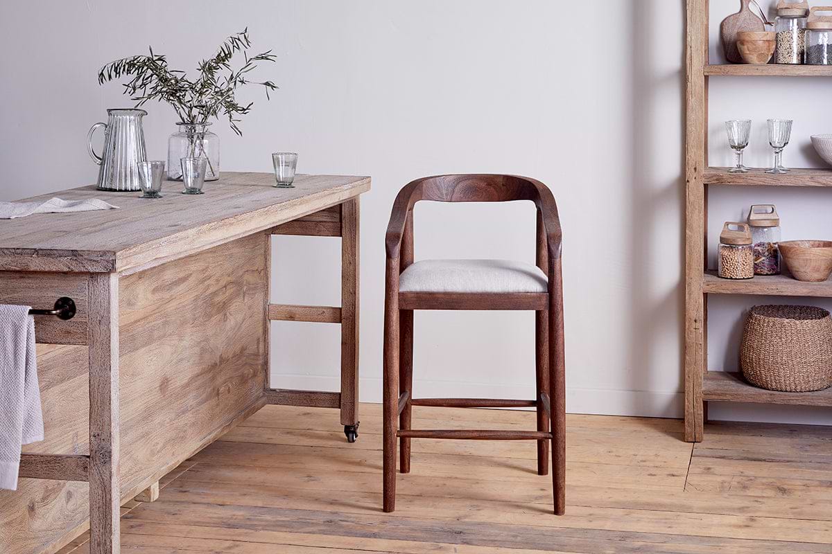 An nkuku Anbu Acacia Upholstered Counter Chair – Washed Walnut with a white cushion sits in a rustic kitchen with light wooden floors, an acacia table on the left with glassware and greenery, and shelves on the right holding baskets, bowls, and kitchen items.