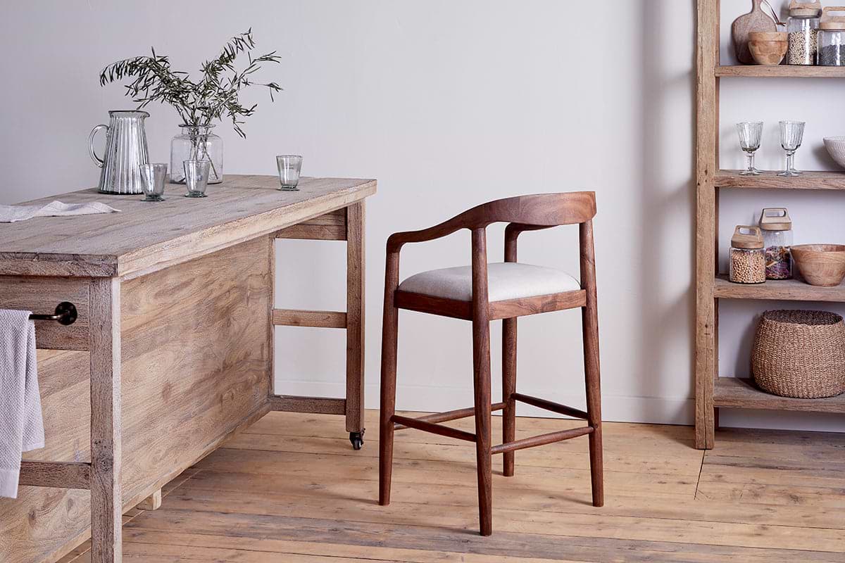 The nkuku Anbu Acacia Upholstered Counter Chair in Washed Walnut, featuring a curved back and light cushion, sits by a rustic acacia table with glassware and foliage, creating a cozy, minimalist space.