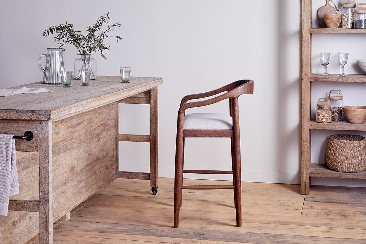 An Anbu Acacia Upholstered Counter Chair by nkuku in washed walnut, with a curved backrest, sits beside a rustic wood table adorned with glassware and greenery. An open wooden shelf holds bowls and jars; acacia floors and neutral walls complete the room.
