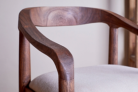 Close-up of the nkuku Anbu Acacia Upholstered Counter Chair - Washed Walnut, showing its curved backrest, armrests, rich brown acacia wood grain, and light beige fabric seat against a softly blurred background.