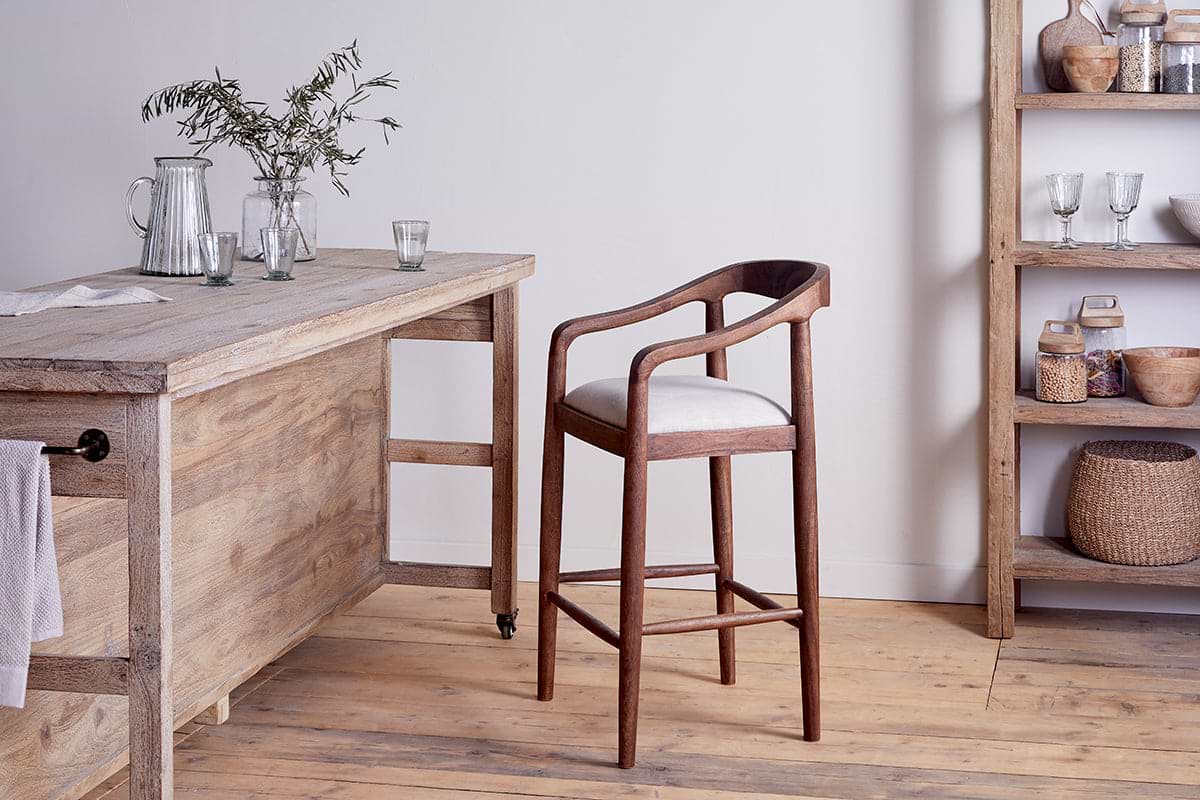 The nkuku Anbu Acacia Upholstered Counter Chair in Washed Walnut, with a white cushion and curved back, pairs with a rustic table amid glass pitchers, cups, and shelves of baskets and jars on a wooden floor.