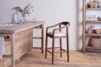 The nkuku Anbu Acacia Upholstered Counter Chair in Washed Walnut, with a white cushion and curved back, pairs with a rustic table amid glass pitchers, cups, and shelves of baskets and jars on a wooden floor.