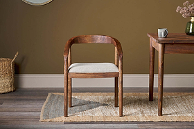 The Anbu Acacia Upholstered Dining Chair by nkuku, in washed walnut with a curved backrest, sits on a tan and gray woven rug beside a wooden table. Brown walls, wood floors, and a wicker basket complete the room, with a ceramic mug and dried flowers on the table.