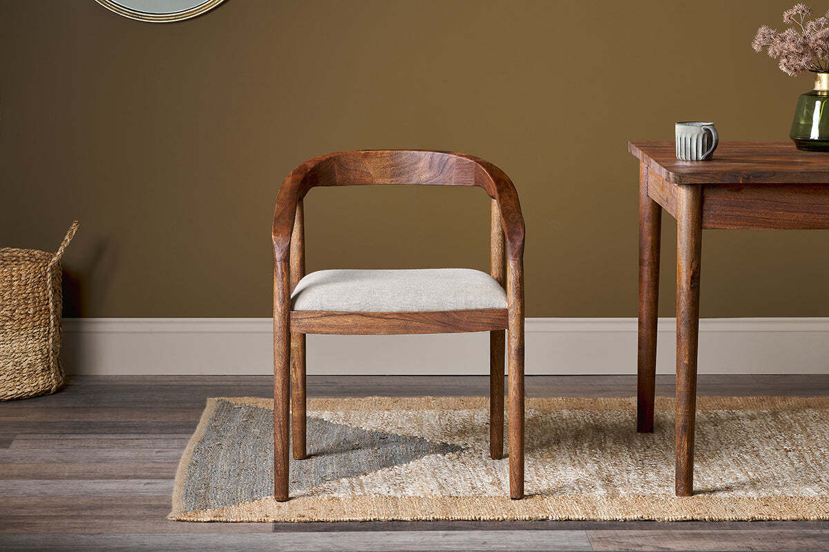 The Anbu Acacia Upholstered Dining Chair by nkuku, in washed walnut with a curved backrest, sits on a tan and gray woven rug beside a wooden table. Brown walls, wood floors, and a wicker basket complete the room, with a ceramic mug and dried flowers on the table.