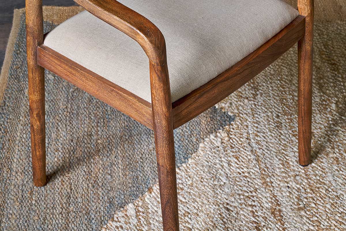 The Anbu Acacia Upholstered Dining Chair by nkuku features curved armrests, a beige seat, and a washed walnut finish. Shown on a woven rug with geometric patterns in natural and gray shades, it highlights smooth wood grain and texture.