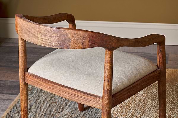 The nkuku Anbu Acacia Upholstered Dining Chair in Washed Walnut features a curved sculpted backrest and armrests with a light beige seat, shown on a textured rug atop wooden flooring against a tan-colored wall.