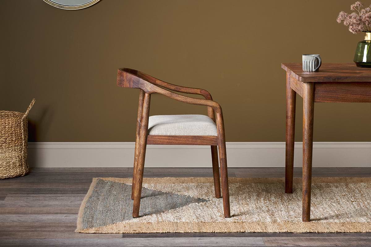 The nkuku Anbu Acacia Upholstered Dining Chair in Washed Walnut, featuring curved arms and a light cushioned seat, sits by a wooden table on a beige and gray rug, with a brown wall, woven basket, ceramic mug and vase in the background.