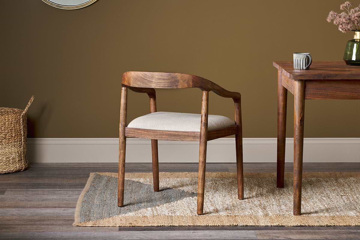 The nkuku Anbu Acacia Upholstered Dining Chair in Washed Walnut, featuring a curved backrest and light beige cushion, sits on a woven rug beside an acacia wood table with a ceramic mug and vase, backed by a brown wall and woven basket.