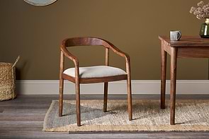 The Anbu Acacia Upholstered Dining Chair by nkuku in washed walnut, featuring a light gray cushion, sits on a patterned rug beside an acacia table, with a woven basket to its left.