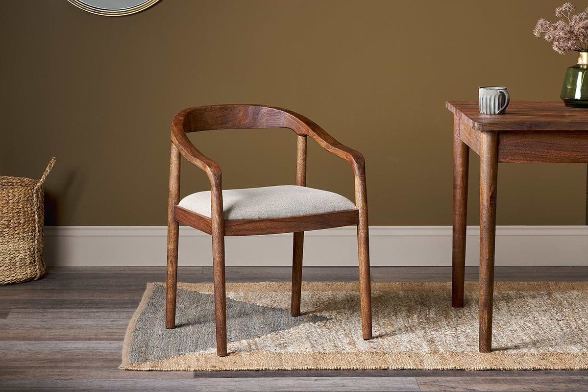 The Anbu Acacia Upholstered Dining Chair by nkuku in washed walnut, featuring a light gray cushion, sits on a patterned rug beside an acacia table, with a woven basket to its left.