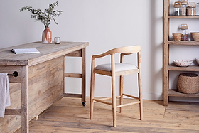 The Anbu Mango Wood Upholstered Counter Chair by nkuku, with a curved backrest and cushioned seat, sits by a rustic wooden table topped with a pink vase of greenery and glassware. Shelves display jars, bowls, and baskets in this minimalist wood-floored room.