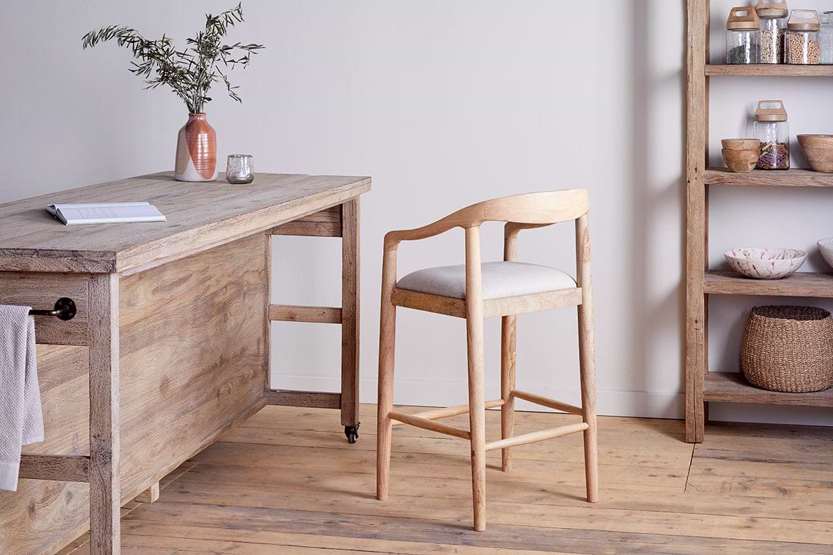 The Anbu Mango Wood Upholstered Counter Chair by nkuku, with a curved backrest and cushioned seat, sits by a rustic wooden table topped with a pink vase of greenery and glassware. Shelves display jars, bowls, and baskets in this minimalist wood-floored room.