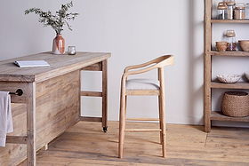 A light wooden desk with a book, candle, and vase stands on a wooden floor. In front is the nkuku Anbu Mango Wood Upholstered Counter Chair - Natural. To the right, shelves hold jars and baskets against a white wall.