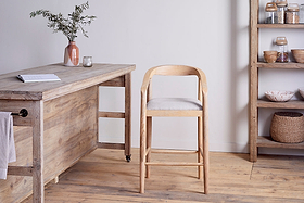 The Anbu Mango Wood Upholstered Counter Chair – Natural by nkuku stands beside a rustic table. A ceramic vase with greenery, a glass, and shelves with jars and baskets complete the minimalist wooden-floored room.