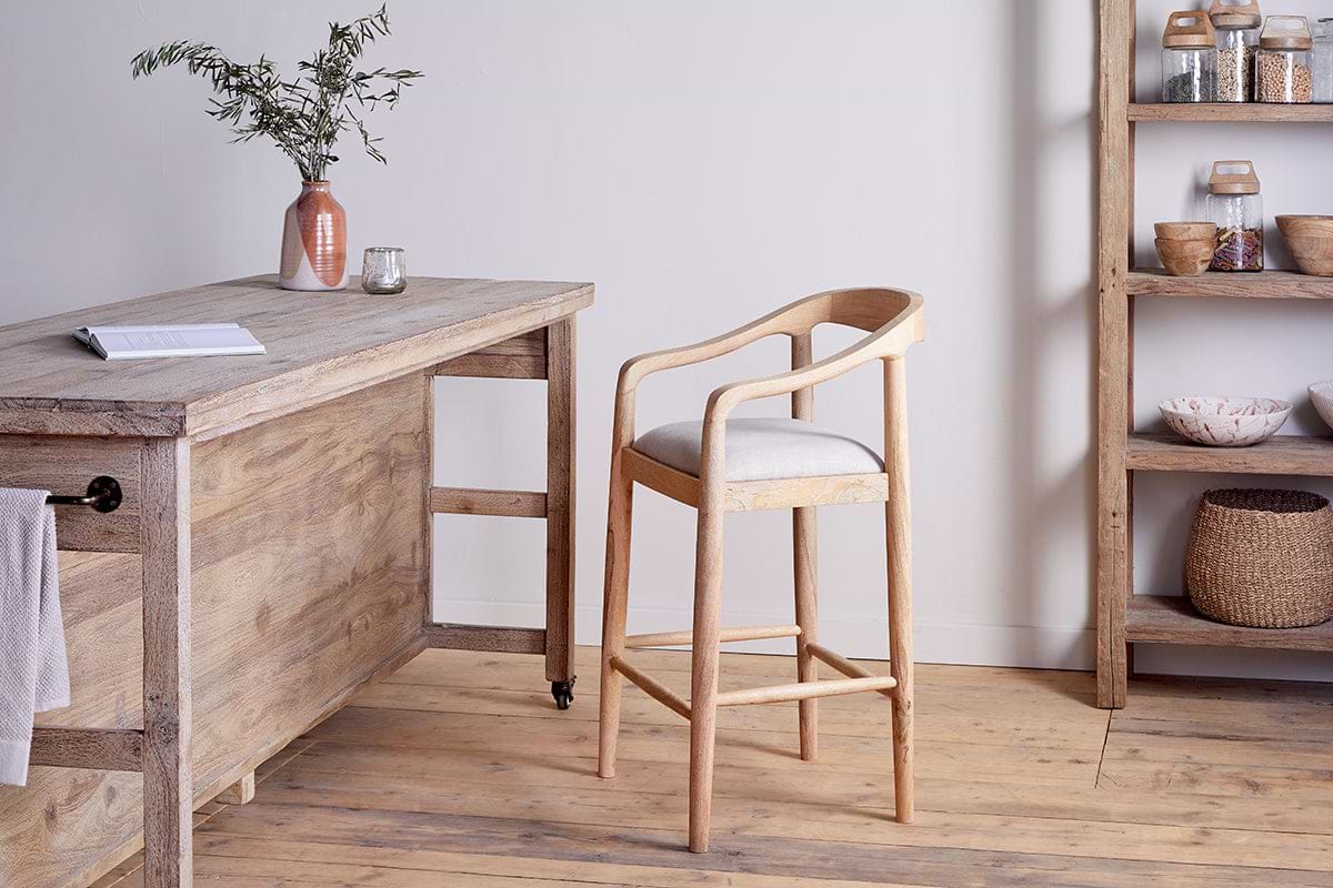 The Anbu Mango Wood Upholstered Counter Chair by nkuku, featuring a curved back and cream cushion, sits beside a rustic table with a pink vase of greenery and candle holder. Shelves holding baskets and jars line the wooden floor.