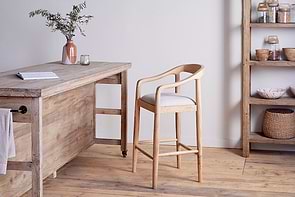 The Anbu Mango Wood Upholstered Counter Chair by nkuku, featuring a curved back and cream cushion, sits beside a rustic table with a pink vase of greenery and candle holder. Shelves holding baskets and jars line the wooden floor.