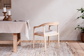The nkuku Anbu Mango Wood Upholstered Dining Chair - Natural, with a curved backrest and cream seat, stands on a neutral woven rug beside a linen-covered table with wooden candleholders. A potted plant and wall art complete the look.