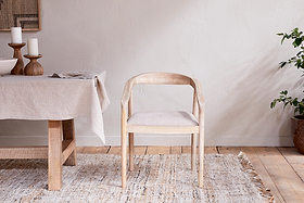 The Anbu Mango Wood Upholstered Dining Chair by nkuku sits on a textured beige rug near a linen-covered dining table with candles, bowls, and napkins. A potted plant decorates the corner against a neutral wall.