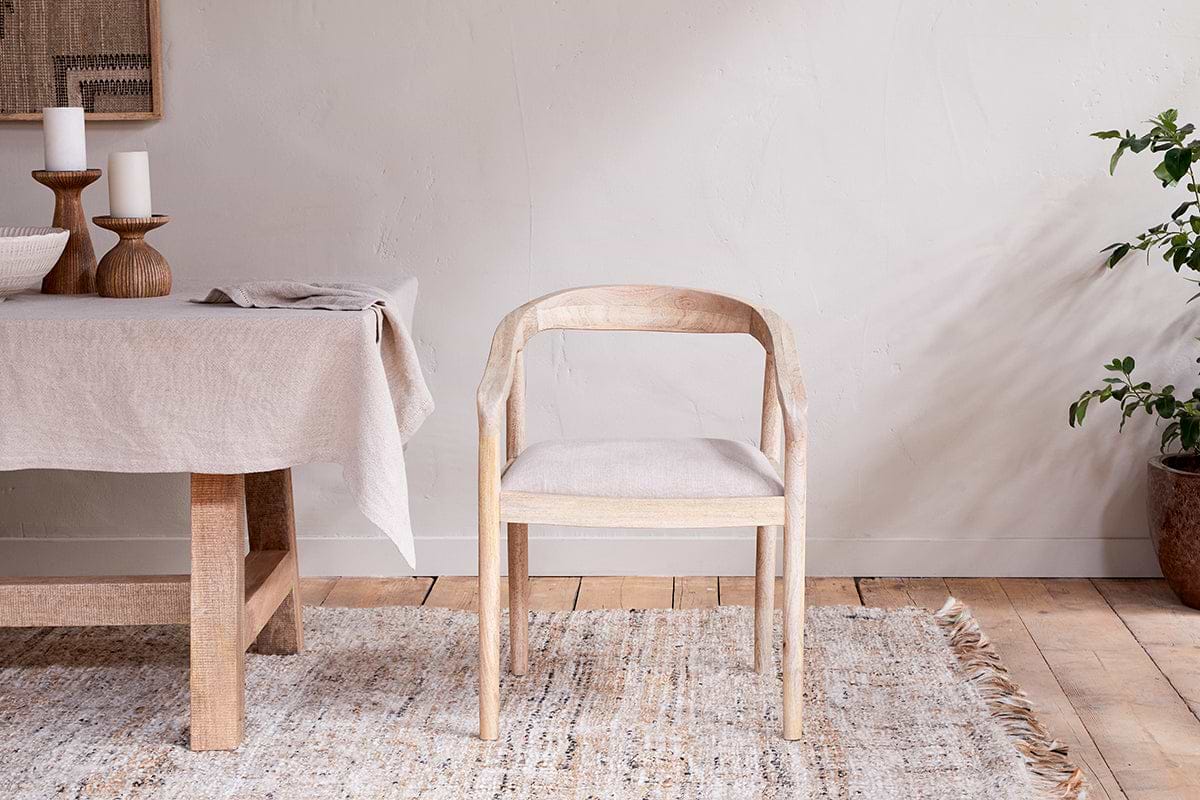 The Anbu Mango Wood Upholstered Dining Chair by nkuku sits on a textured beige rug near a linen-covered dining table with candles, bowls, and napkins. A potted plant decorates the corner against a neutral wall.