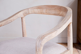 Close-up of the nkuku Anbu Mango Wood Upholstered Dining Chair - Natural, featuring a smooth light wood finish, elegantly curved backrest, armrests, and a light beige padded seat set against an off-white wall.