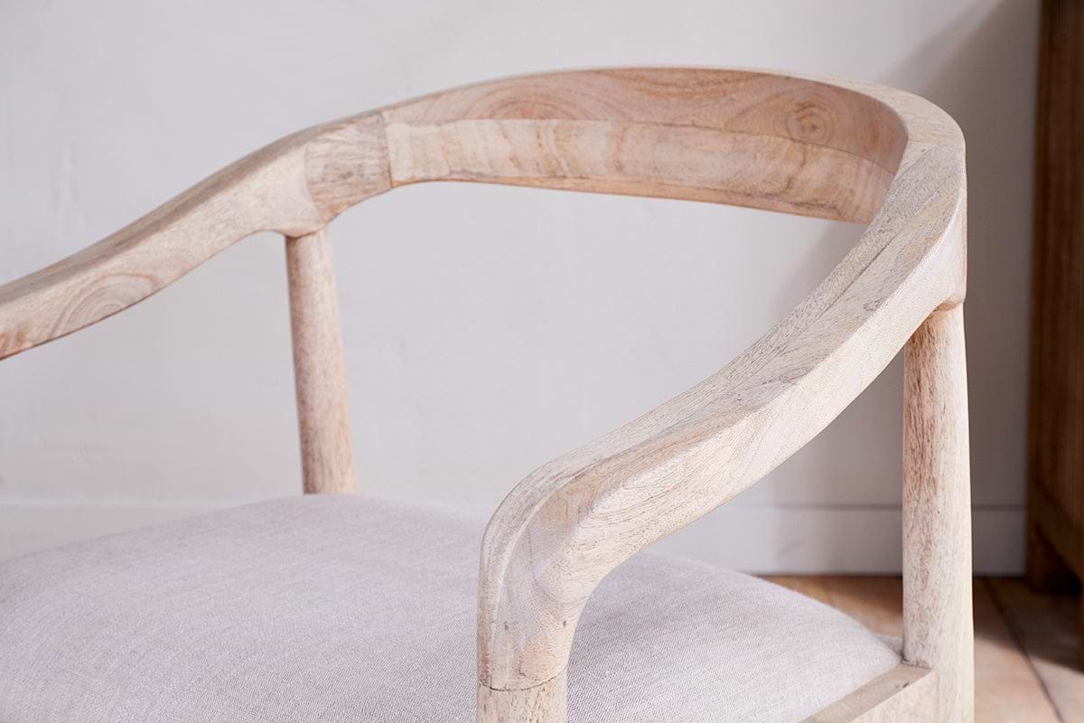 Close-up of the nkuku Anbu Mango Wood Upholstered Dining Chair - Natural, featuring a smooth light wood finish, elegantly curved backrest, armrests, and a light beige padded seat set against an off-white wall.