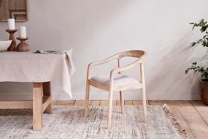 The Anbu Mango Wood Upholstered Dining Chair by nkuku features curved armrests and a cream seat, sitting on a neutral woven rug beside a linen-covered table with candles. A potted plant adds greenery.